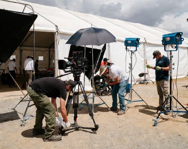 Film crew cleaning and preparing an outdoor movie set with lighting equipment visible